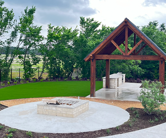 Outdoor pavilion featuring a stone fire pit and manicured lawn area surrounded by greenery and landscaping.