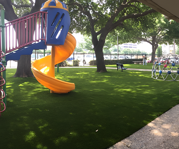 Bright orange slide and climbing dome in a playground with artificial grass, surrounded by trees and open space.